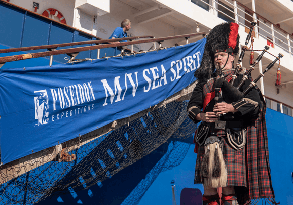 Vor dem Expeditionskreuzfahrtschiff steht ein Mann mit einem Dudelsack und begrüsst die Kreuzfahrtgäste vor der Abfahrt.
